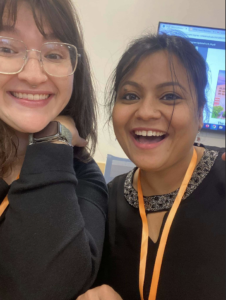A close up of Emily and Meghalee smiling at a camera with a large screen in the background. Both are wearing black with their conference lanyards around their necks.